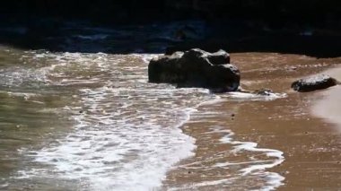 Küçük dalgalar yavaşça sahile doğru hareket ediyor ve kumlara saplanmış bir kayaya doğru..