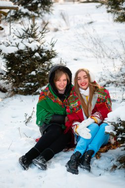 Two Ukrainian sisters are walking in the park in winter, smiling and wearing warm, beautiful clothes. Many Christmas trees around