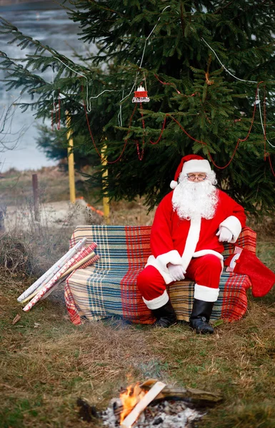 Santa Claus is sitting in nature by the fire near the Christmas trees. Red suit with a white beard. Winter holidays in the world