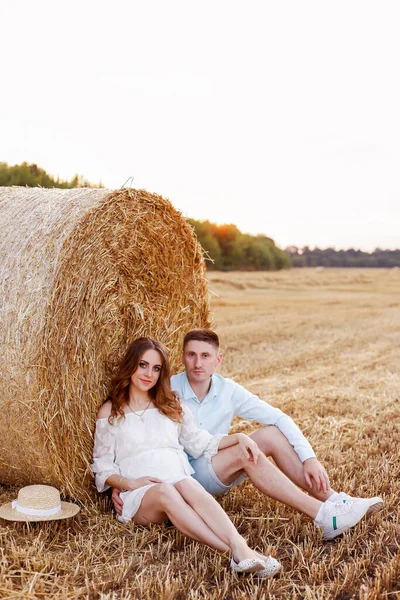 A pregnant woman with her husband on a summer walk in a wheat field. Fashionable summer clothes. White dress