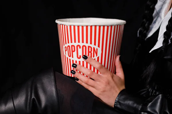 A brunette with two pigtails in a black dress holds a box of popcorn. Wednesday series from Netflix. Copy space
