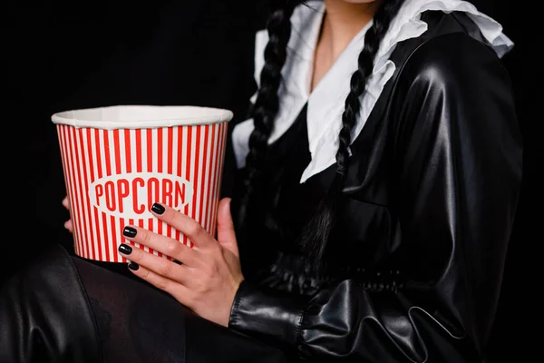 A brunette with two pigtails in a black dress holds a box of popcorn. Wednesday series from Netflix