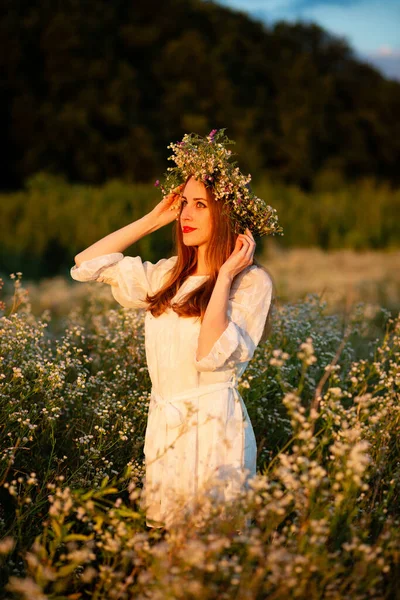 A girl with a smile stands in a field with a wreath on her head at sunset in a white dress