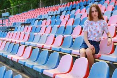 The girl sits on the benches at the stadium and watches football on the grass. She is worried about the game