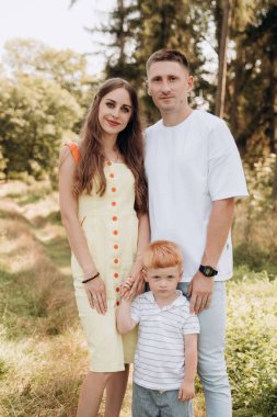 A young family walks in a coniferous park and spends time happily together. A young family walks in a coniferous park and spends time happily together. Mom is in a yellow dress, and dad and son are in white T-shirts