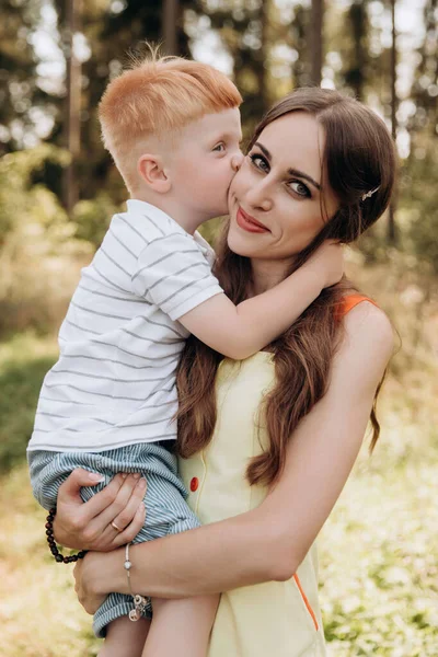 A red-haired son kisses his mother on the cheek. Family values are the most precious. Coniferous forest on the background