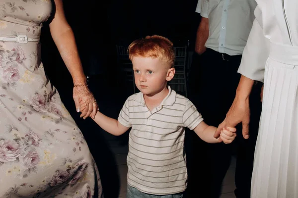 A red-haired boy holds the hands of two grandmothers while dancing at a wedding