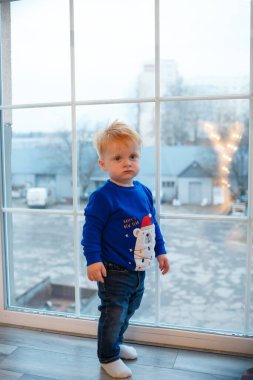 Little red-haired boy 2 years old in a New Year's sweater near the big window at home. Waiting for Christmas