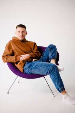 Portrait of a young 30-year-old man. Brown sweatshirt and blue jeans. Purple stylish chair. Short hairstyle