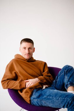 Portrait of a young 30-year-old man. Brown sweatshirt and blue jeans. Purple stylish chair