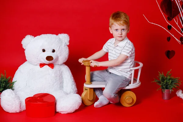 A little boy with a white bear on a red background. Children's white wooden bicycle