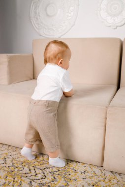 1 yaşında bir çocuk, evinde, oturma odasında, beyaz bir kanepenin yanında oynuyor.