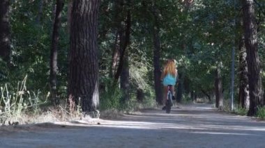 Bir kız geniş bir park yolu boyunca bisiklet sürüyor. Omuzlarında uzun kıvırcık saçlar dalgalanıyor. Dağ bisikletleri için orman patikası. Yüksek kalite 4k görüntü