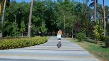 Sarışın kız bir şehir parkında dağ bisikleti üzerinde yürüyor. Bir kız, geniş lastikleri ve lastikleri olan yeşil bir çiçek tarhında bisiklet sürüyor. Yüksek kalite 4k görüntü