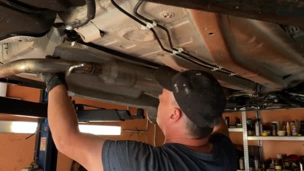 一个工匠在他的车间里拆车 汽车维修工人拆卸汽车的排气系统 修理一辆凸起的汽车 从下面看一位机械师修理汽车底部 — 图库视频影像