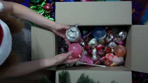 赤い帽子をかぶった若い女性は クリスマスツリーのおもちゃを選びます 箱の中にあるクリスマスツリーの装飾 新年の休日に 少女は彼女が選ぶ古いおもちゃでクリスマスツリーを飾ります — ストック動画
