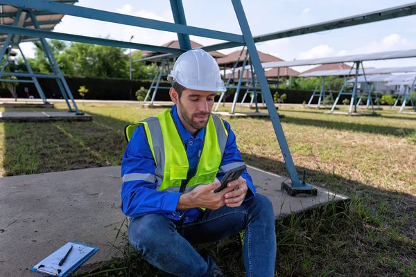 休憩時間に太陽光発電所のエンジニアは 太陽光パネルの下で携帯電話からソーシャルメディアを見て座って — ストック写真