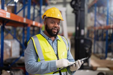 Depoda çalışan Afrikalı Amerikalı forkliftleri kontrol ediyor, gülümsüyor.
