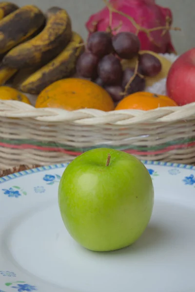 green apples on a white classic plate with a basket of fruit in the background.