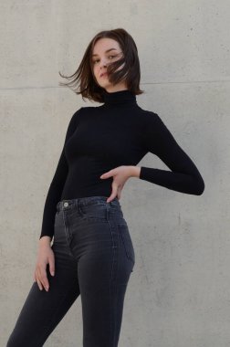Genç moda kızı gri beton bir duvarın önünde kot pantolon ve siyah bir kazak giyiyor..