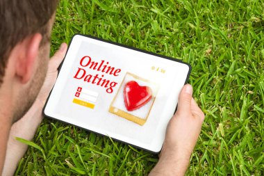 Young man lying on the grass holding a tablet device and visiting an online dating website. clipart