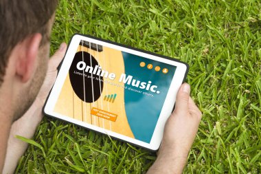 Bir internet sitesini ziyaret eden genç adam tablet cihazıyla çimlerin üzerinde yatıyor..