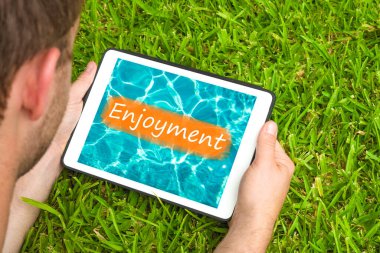 Young man lying on grass in garden with tablet device looking at water background with word 