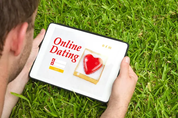 Genç bir adam elinde tablet cihazıyla çimlerin üzerinde uzanıyor ve bir arkadaşlık sitesini ziyaret ediyor..