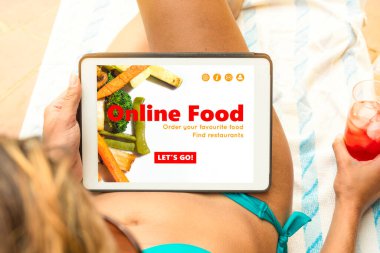 Bikinili genç bir kadın tablet cihazıyla online yemek sitesini ziyaret ediyor, plaj havlusuna uzanıyor ve bir şeyler içiyor..