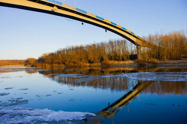 a bridge across the river with reflection in the water as well as trees on the shore and ice in the water in the rays of the warm setting sun in winter in Europe in Ukraine in Chernihiv