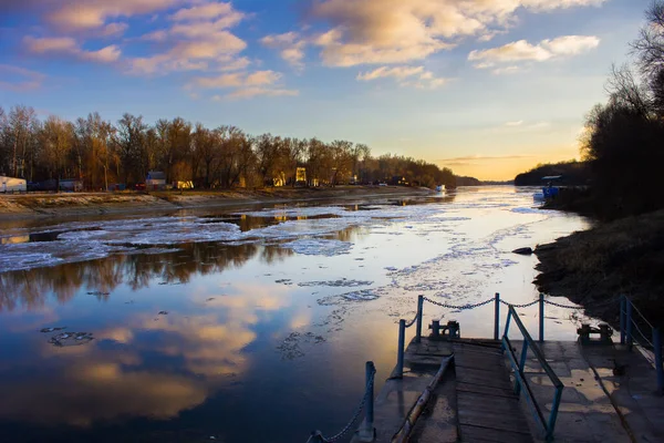 a river with a reflection in the water as well as trees on the shore and ice in the water in the rays of a warm setting sun in winter, as well as a pier on the shore and texture clouds in the sky in Europe in Ukraine