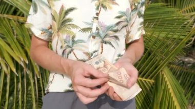 Güneşli bir günde arkada palmiye yaprakları olan tropikal gömlekli genç bir kadın Tayland Bahti 'ni sayıyor. Kapat..