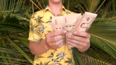 Siyah güneş gözlüklü ve sarı tropikal tişörtlü genç bir adam güneşli bir günde Tayland 'da bir tropik cennet adasında arka planda palmiye yaprakları olan bir plajda Tayland Bahti' ni sayıyor..