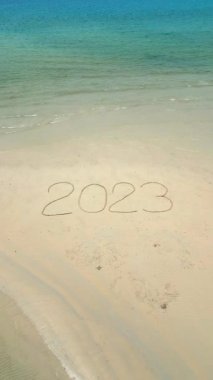 Turkuaz okyanus suyuyla çevrili tropikal bir sahildeki beyaz kumun üzerine yazılmış 2023 sözcüğünün havadan görünüşü..