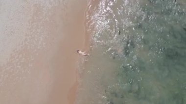 Dalgalar zarif bir şekilde ona doğru süzülürken, bikinili genç bir kadının ışıl ışıl denizin kenarında güneşlenişi..