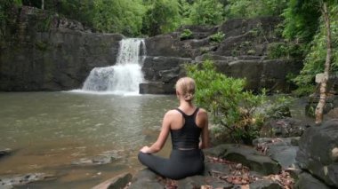 Genç ve güzel bir kadın Tayland 'daki tropikal yağmur ormanlarında inanılmaz bir şelalede meditasyon yapıyor..