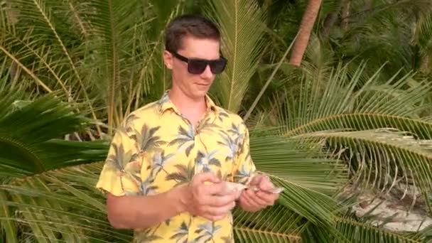 在泰国的一个阳光明媚的日子里 一个戴着黑色太阳镜 身穿黄色热带衬衫的年轻人正在数着背景上有棕榈叶的泰铢 — 图库视频影像