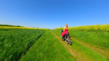 Kırmızı elbiseli beyaz bir kadın ve kırsalda klasik bir motosiklete binen bir köpek ağır çekimde sarı kolza tohumu tarlalarından geçiyor.