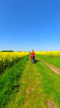 Beyaz bir kadın, kırsalda eski model bir motosiklet sürüyor. Yavaş çekimde sarı kolza tohumu tarlalarından geçiyor..