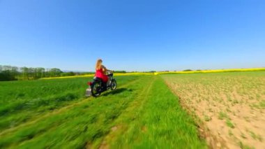 Beyaz kadın ve köpek FPV kırsalda sarı çiçekli tarlalarda klasik bir motosiklet sürüyor.
