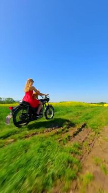 Beyaz kadın ve köpek FPV kırsalda sarı çiçekli tarlalarda klasik bir motosiklet sürüyor.