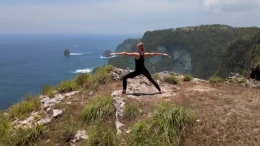 Savaşçı pozuyla yoga yapan atletik bir kadının okyanusun üzerindeki bir uçurumda duruşu. Aktif yaşam tarzı, farkındalık, sağlıklı yaşam kavramları