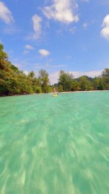 Tayland 'da tropik bir adada kristal berrak denizde kürek çeken bir kadının FPV görüntüsü.
