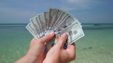 Tropikal plajlarda, zenginlik, seyahat ve finansal özgürlük kavramında Amerikan dolarlarını tutan ellerin yakın çekimi