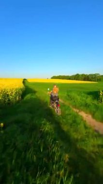 Güneşli bir bahar gününde sarı kolza tohumu tarlalarında motosiklet süren bir kadının FPV drone görüntüsü. Manzaralı kırsal alan.