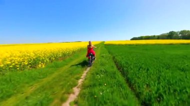 Kırmızı elbiseli bir kadının sarı kolza tohumu tarlalarında motosiklet sürerken çekilmiş dinamik FPV görüntüsü. Özgürlük, bahar ve doğa kavramı.