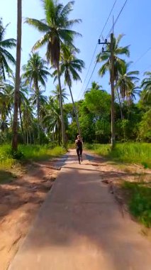 Güneşli bir günde yeşil palmiye ağaçlarıyla tropikal yolda koşan bir kadının sinematik FPV görüntüsü. Egzotik doğada sağlıklı bir yaşam tarzı.