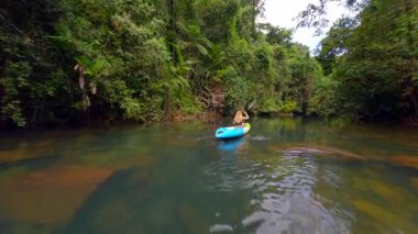 FPV 'de bir kadının yemyeşil ormanda tropikal nehirde kano sürerken görüntüsü. Huzurlu, manzaralı doğa görüntüleri seyahat, macera ve eko-turizm projeleri için mükemmeldir..
