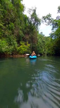 FPV 'de bir kadının yemyeşil ormanda tropikal nehirde kano sürerken görüntüsü. Huzurlu, manzaralı doğa görüntüleri seyahat, macera ve eko-turizm projeleri için mükemmeldir..