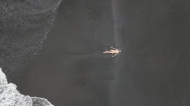 Sarı mayo giymiş, volkanik siyah kumsalda uzanan ve Endonezya, Bali 'de okyanus dalgaları olan bir kadın.